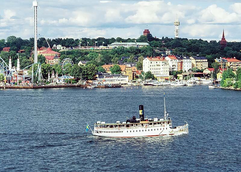 ספינה קטנה לבנה עם ארובה שחורה מפליגה על אגם, ברקע שטוקהולם