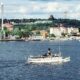 ספינה קטנה לבנה עם ארובה שחורה מפליגה על אגם, ברקע שטוקהולם