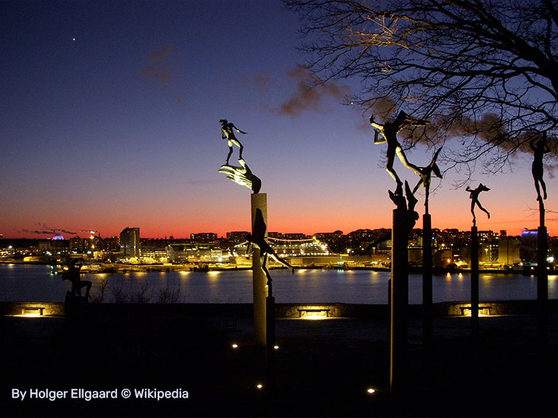 תמונת ערב של גן הפסלים, הפסלים מוארים וברקע העיר שטוקהולם בשקיעה
